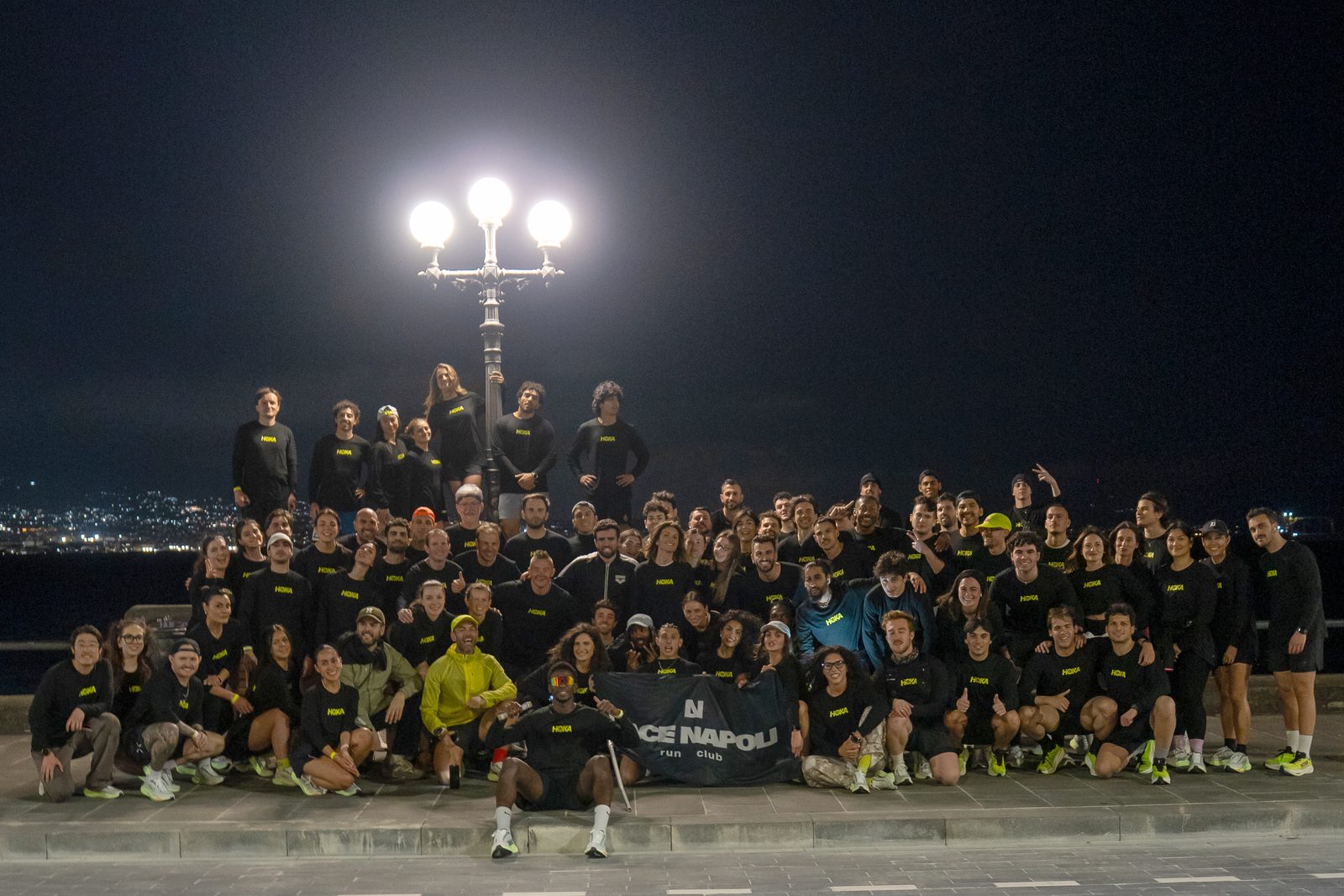 The whole group in black HOKA long sleeves under a street lamp on the Naples seafront at night, holding the LACE Napoli run club banner