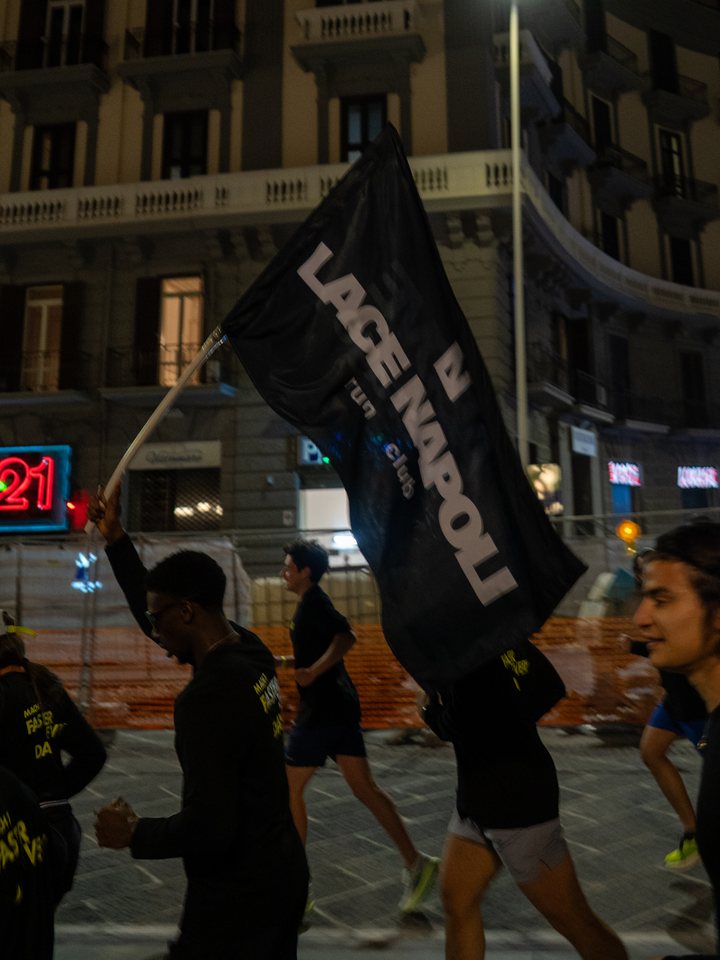 A runner carrying the LACE Napoli run club flag through the streets of Naples at night, with the group running alongside